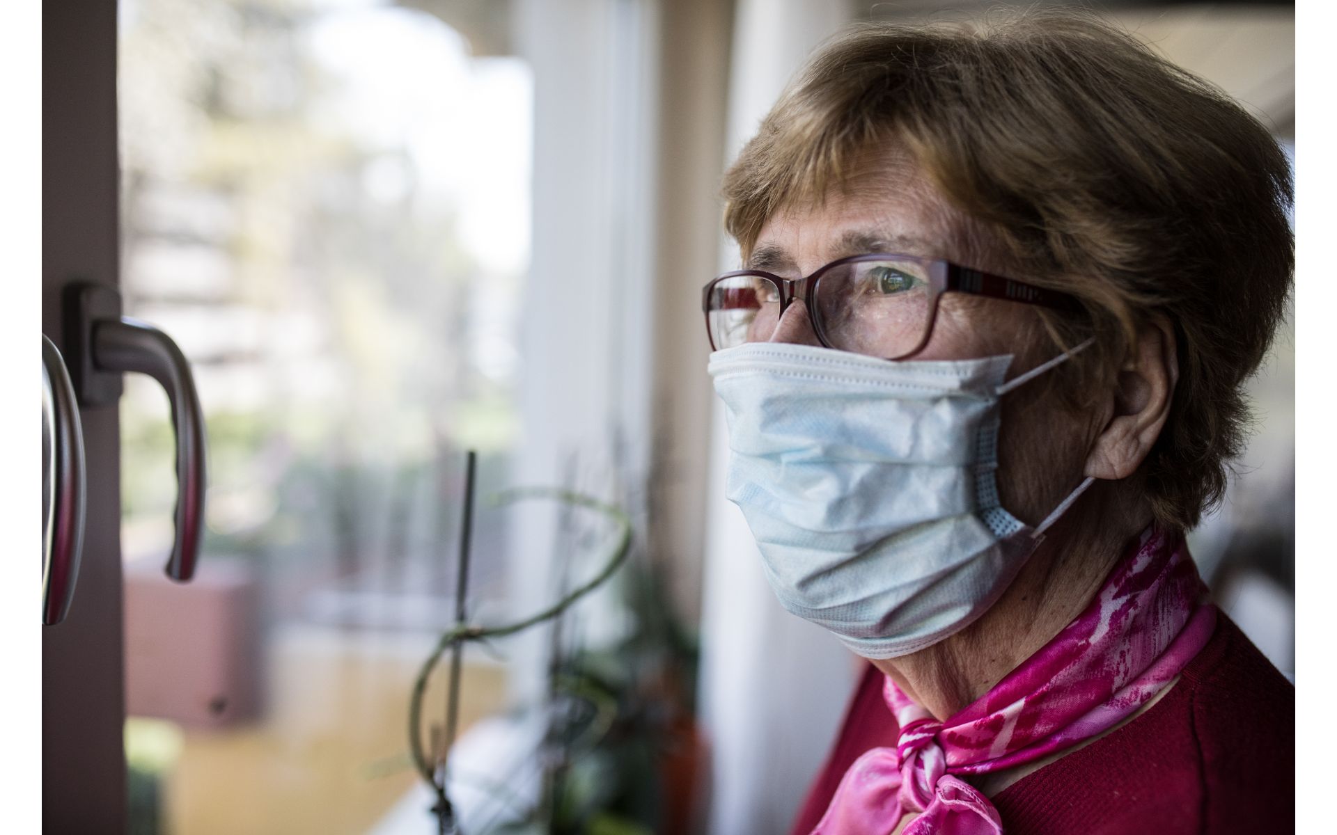 Older woman looks out the window of home while wearing mask.