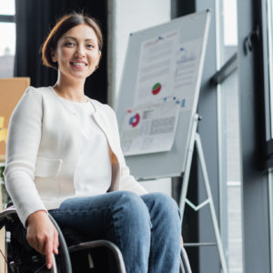 cheerful businesswoman in wheelchair looking at camera near blurred infographics in office