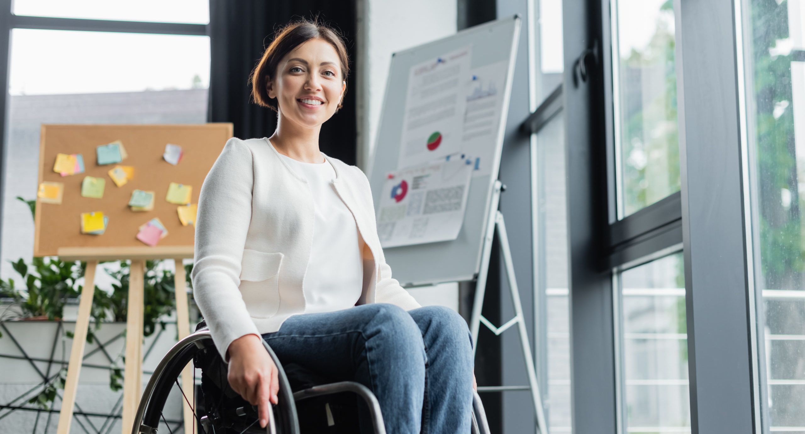 cheerful businesswoman in wheelchair looking at camera near blurred infographics in office
