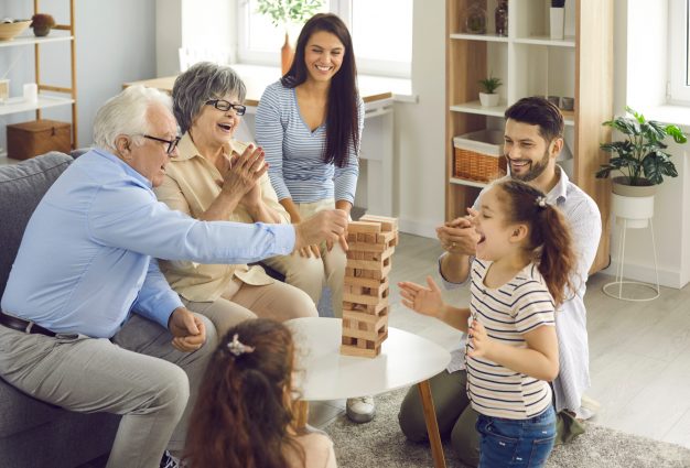 Happy big family with little children enjoying the weekend at home, playing board games and having a good time together. Excited grandkids having fun watching grandpa take a wood block from the tower